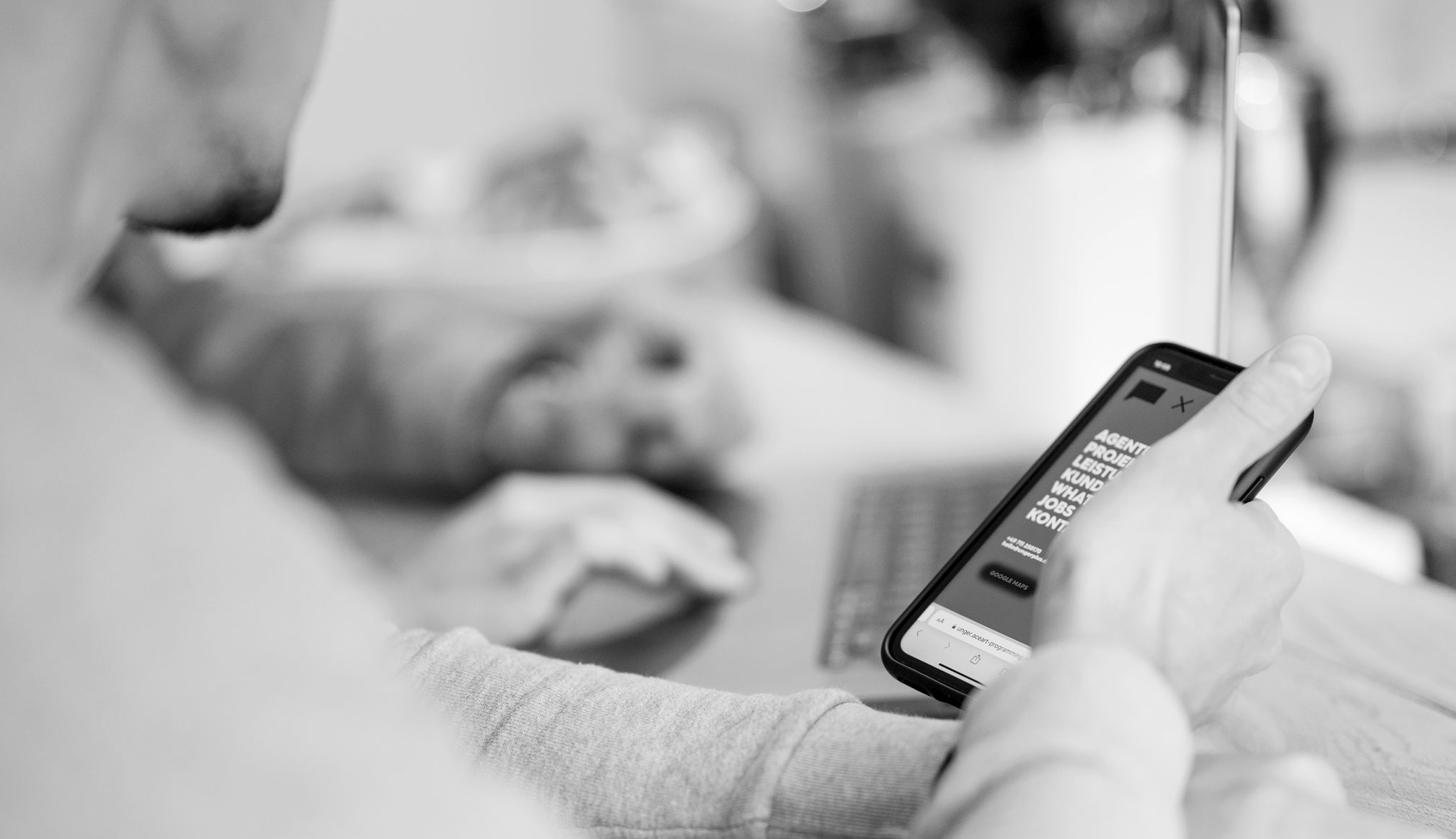 Person hält ein Smartphone mit der mobilen Website von ungerplus, im Hintergrund ein Laptop